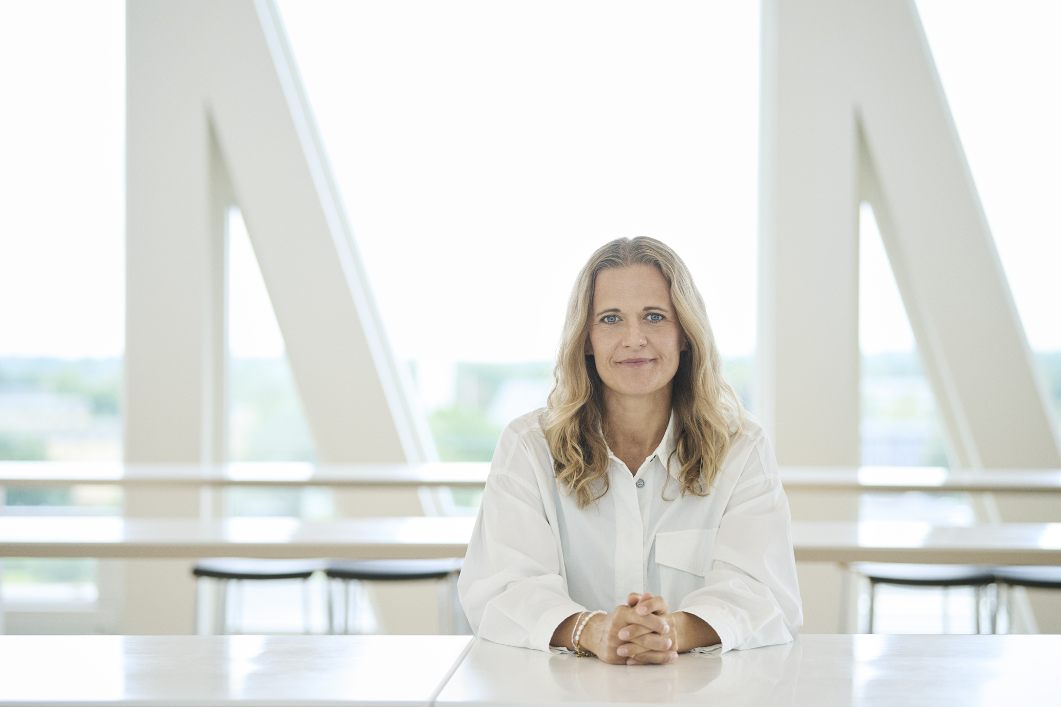 A blonde woman in a white shirt sits at a table with hands folded