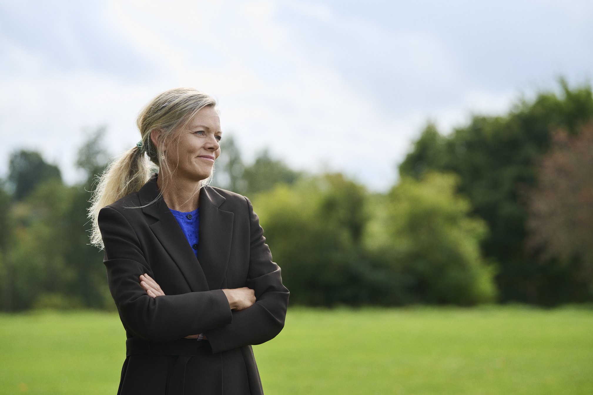 A woman stands outside in a dark professional jacket with her arms crossed 